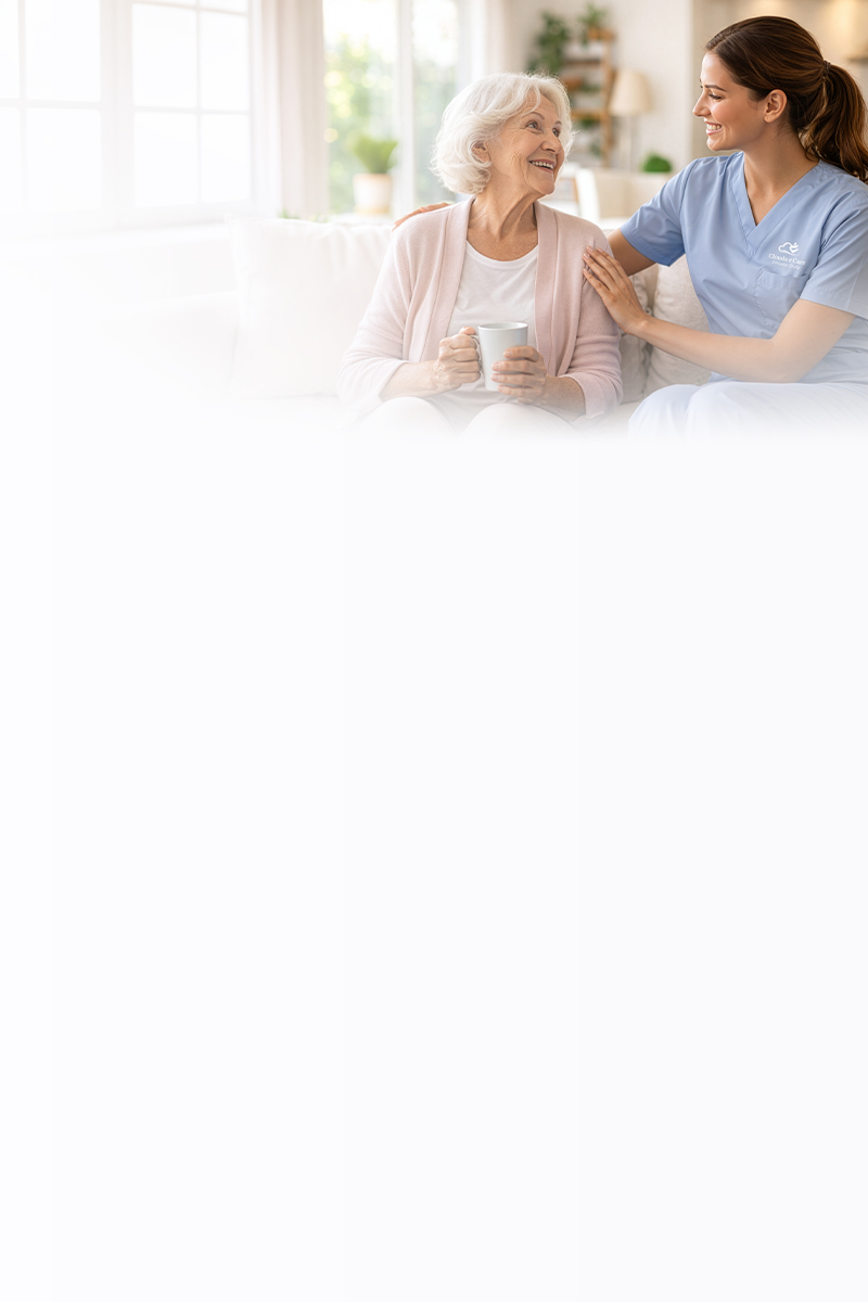 A Clouds of Care caregiver in blue scrubs sits with an elderly woman on a couch, sharing a warm moment at home