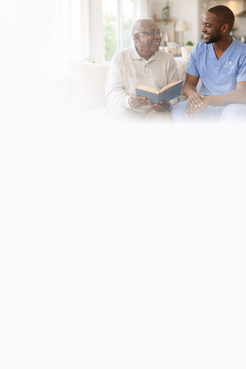 A Clouds of Care caregiver in blue scrubs sits with an elderly man on a couch, sharing a laugh while reading a book together at home