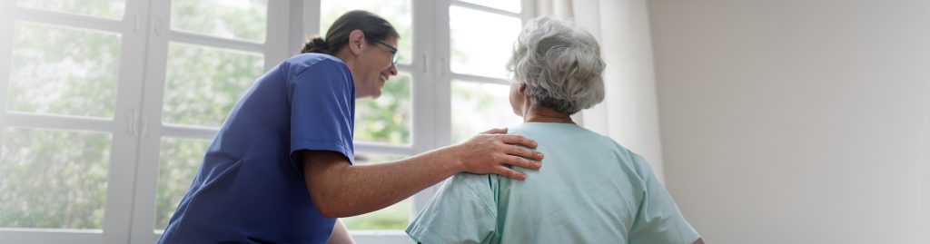 Professional caregiver smiling and placing a supportive hand on the shoulder of an elderly woman with dementia at home
