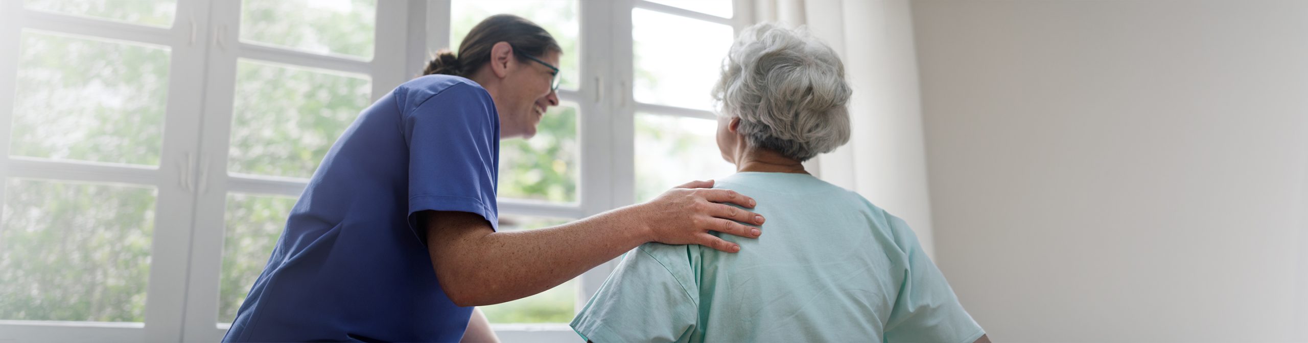 Professional caregiver smiling and placing a supportive hand on the shoulder of an elderly woman with dementia at home