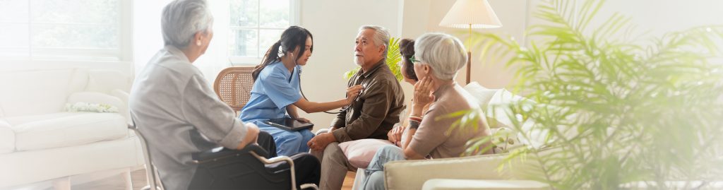 Professional caregiver using a stethoscope to assess a senior man at home while family members sit nearby