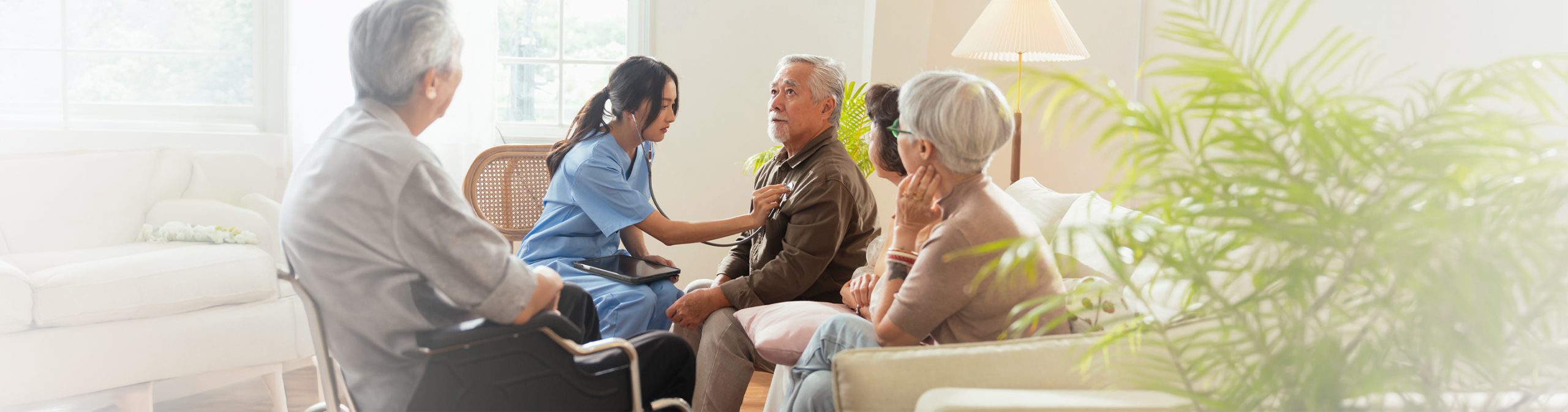Professional caregiver using a stethoscope to assess a senior man at home while family members sit nearby